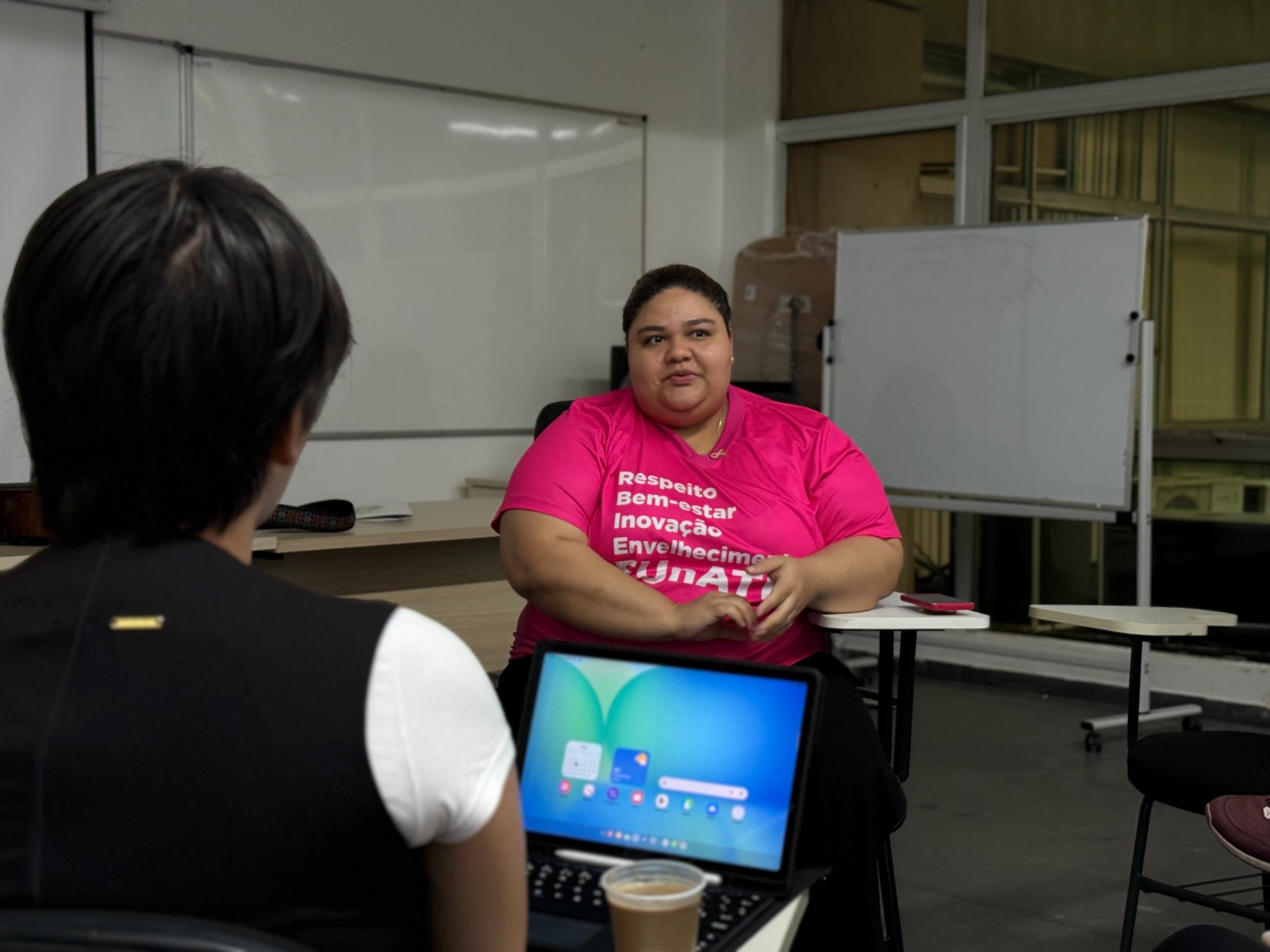 Foto mostra uma pessoa do sexo feminino, sentada, e vestida com camisa rosa, tendo à frente uma pessoa de costas
