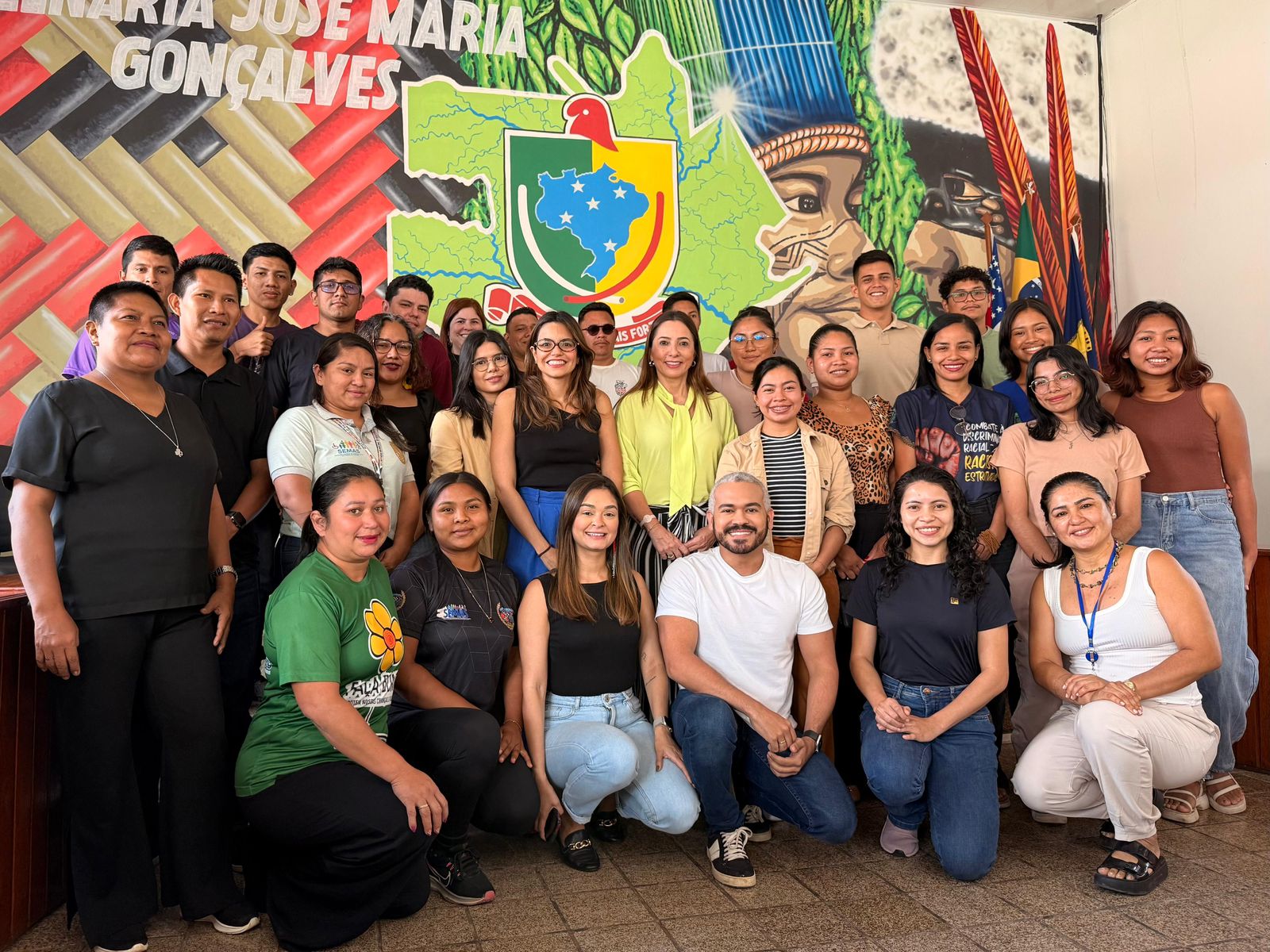 Foto traz diversas pessoas em frente a um mural indígena