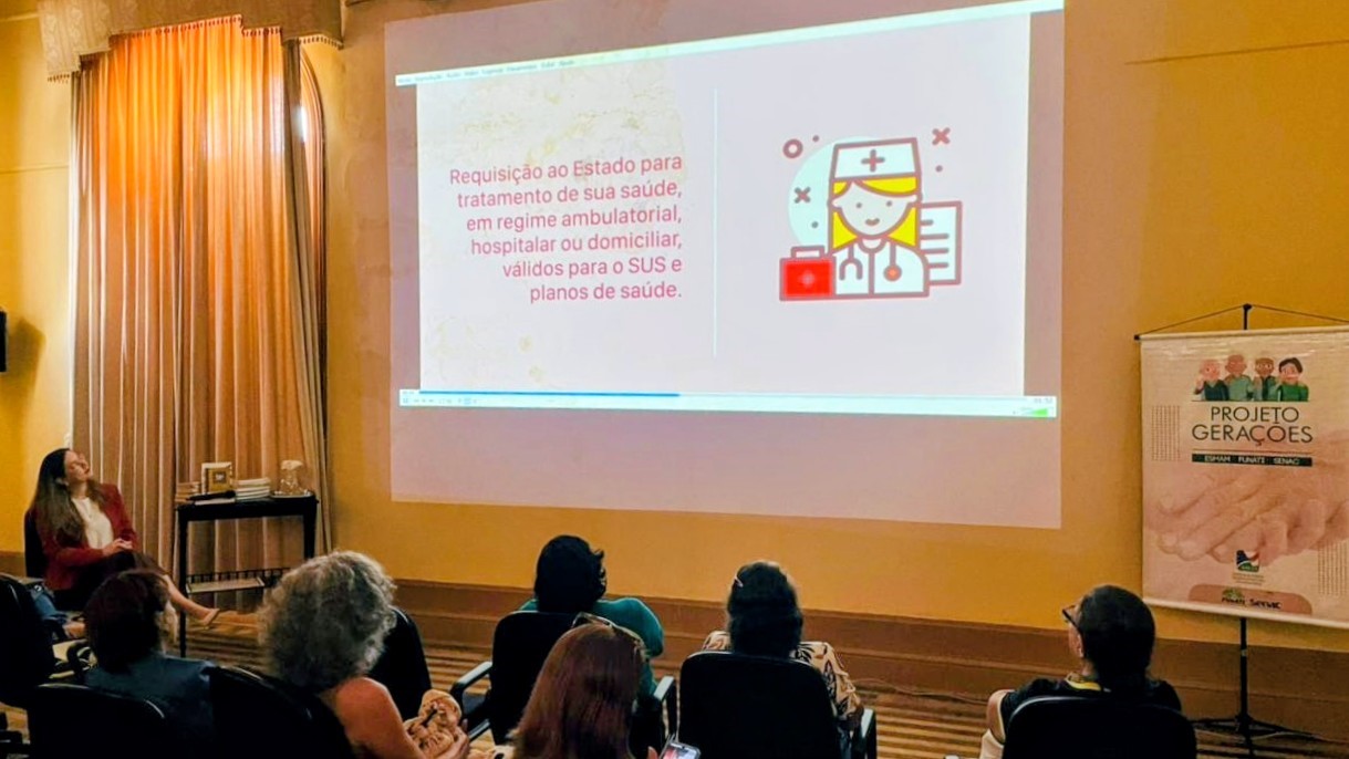 Foto mostra pessoas, sentadas, olhando em direção a um telão