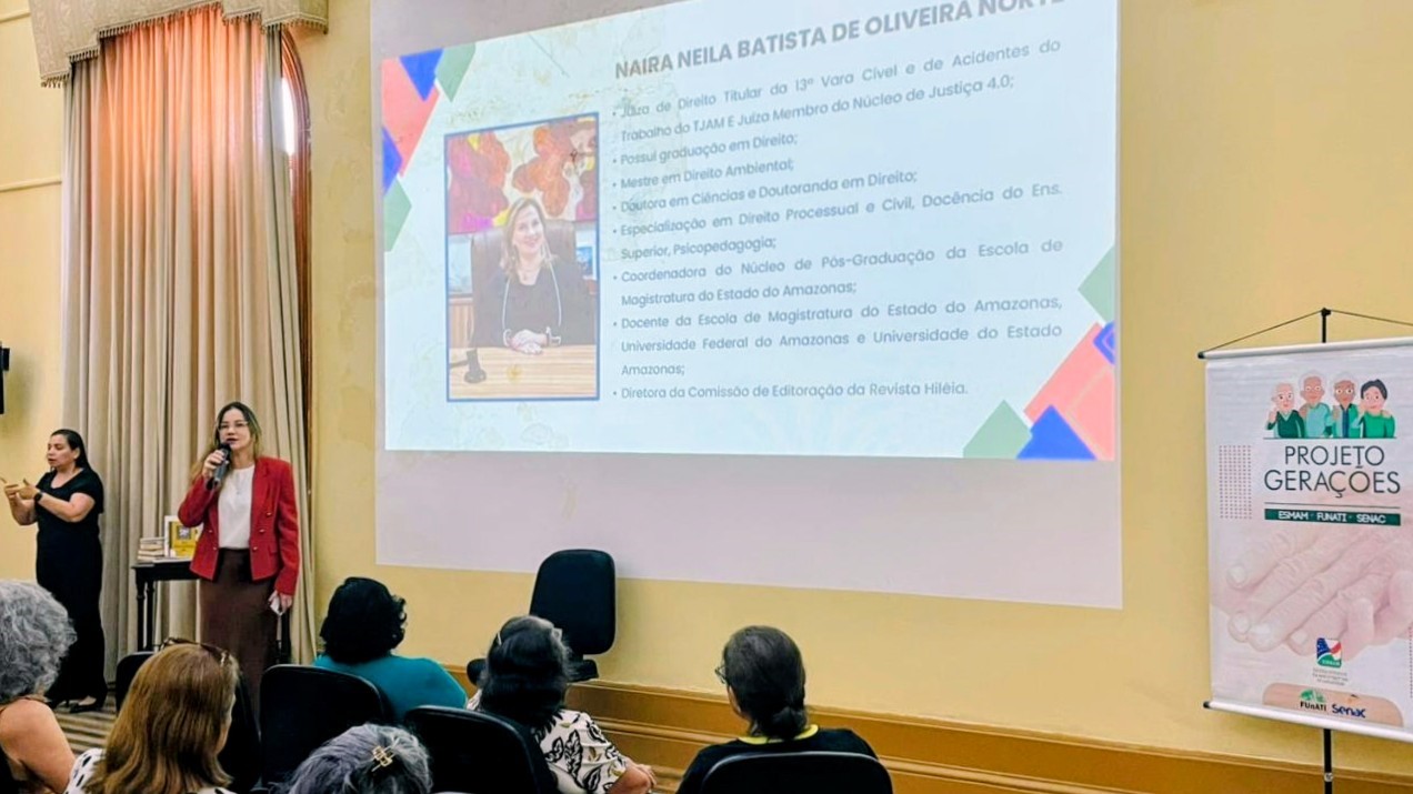 Foto mostra duas pessoas se comunicando com o público, ambas ao lado de um telão